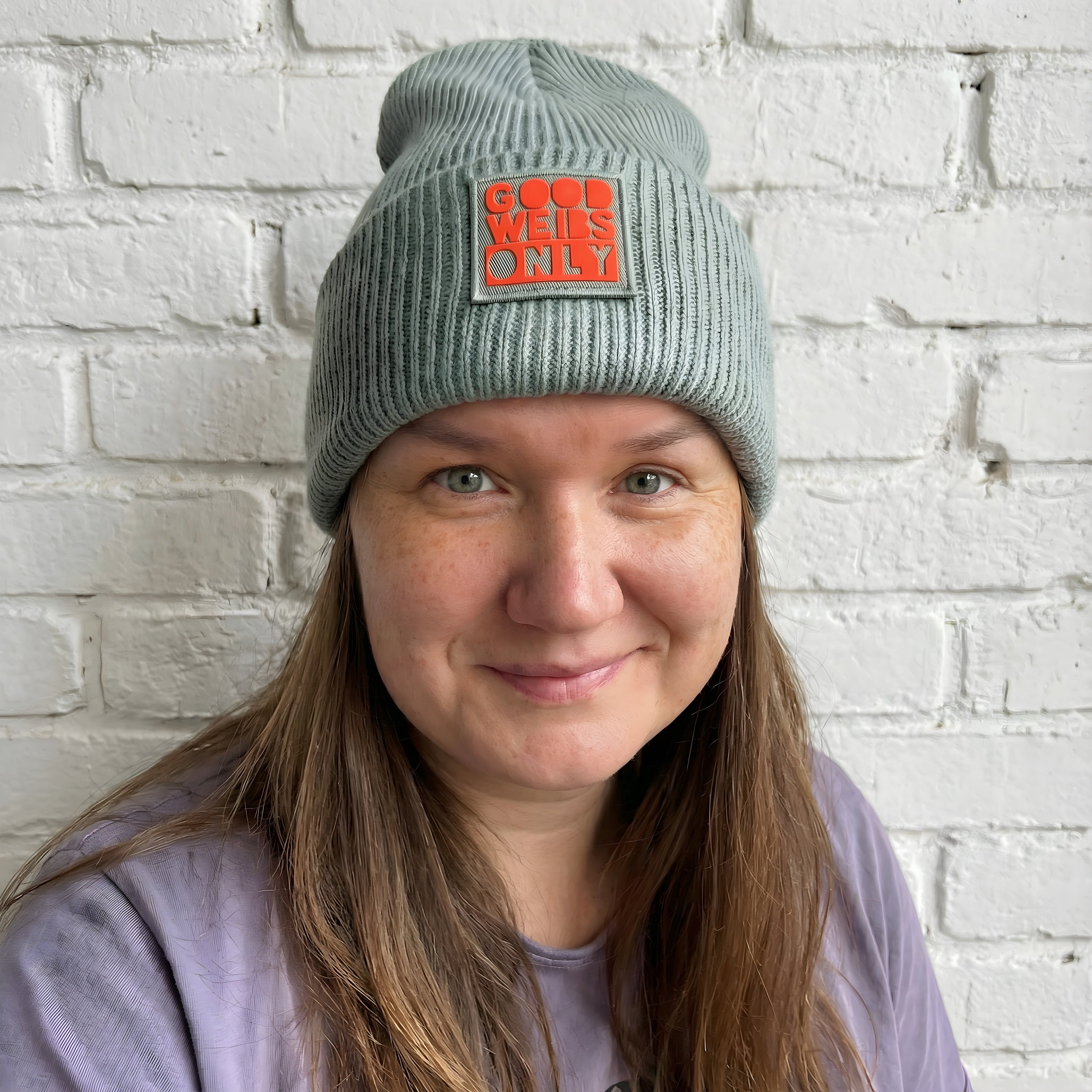Frau trägt grau-grüne Beanie Strickmütze mit rotem Patch ‚Good Weibs Only‘ – handveredeltes TinyTypo-Winteraccessoire.