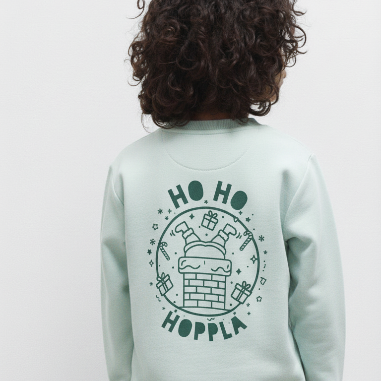 Kind von hinten in mintgrünem Sweatshirt mit Weihnachtsdruck „Ho Ho Hoppla“, Santa steckt kopfüber im Kamin, dunkler Siebdruck.
