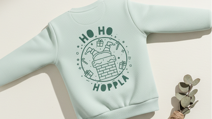 Mintgrünes Kinder-Sweatshirt mit „Ho Ho Hoppla“ Weihnachtsmotiv als Flatlay auf hellem Hintergrund, daneben ein kleiner Eukalyptuszweig.