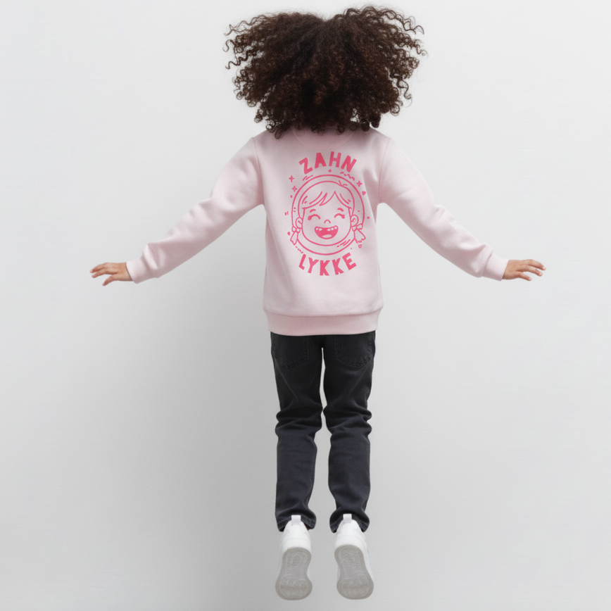 Kind springt, von hinten, trägt soft pink Kinder-Sweatshirt mit „Zahn Lykke“ Motiv, dunkle Jeans und weiße Sneaker