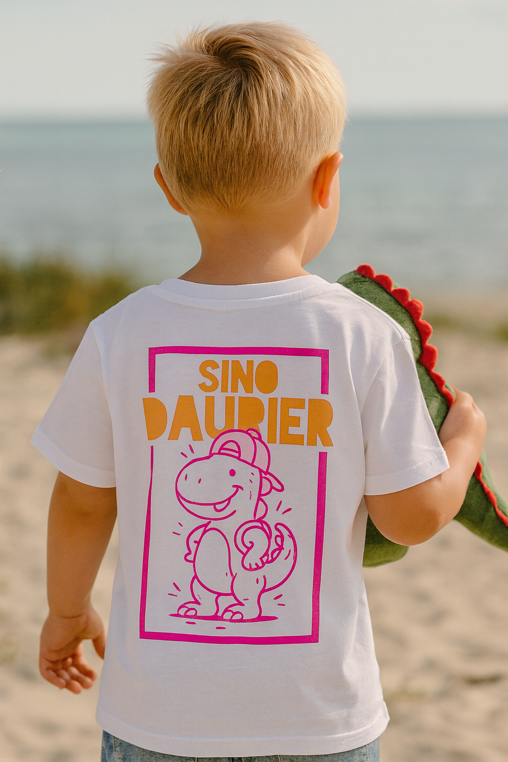 „Kind trägt weißes Tiny-Typo T-Shirt mit SinoDaurier-Design am Strand – handbedruckt, nachhaltig.