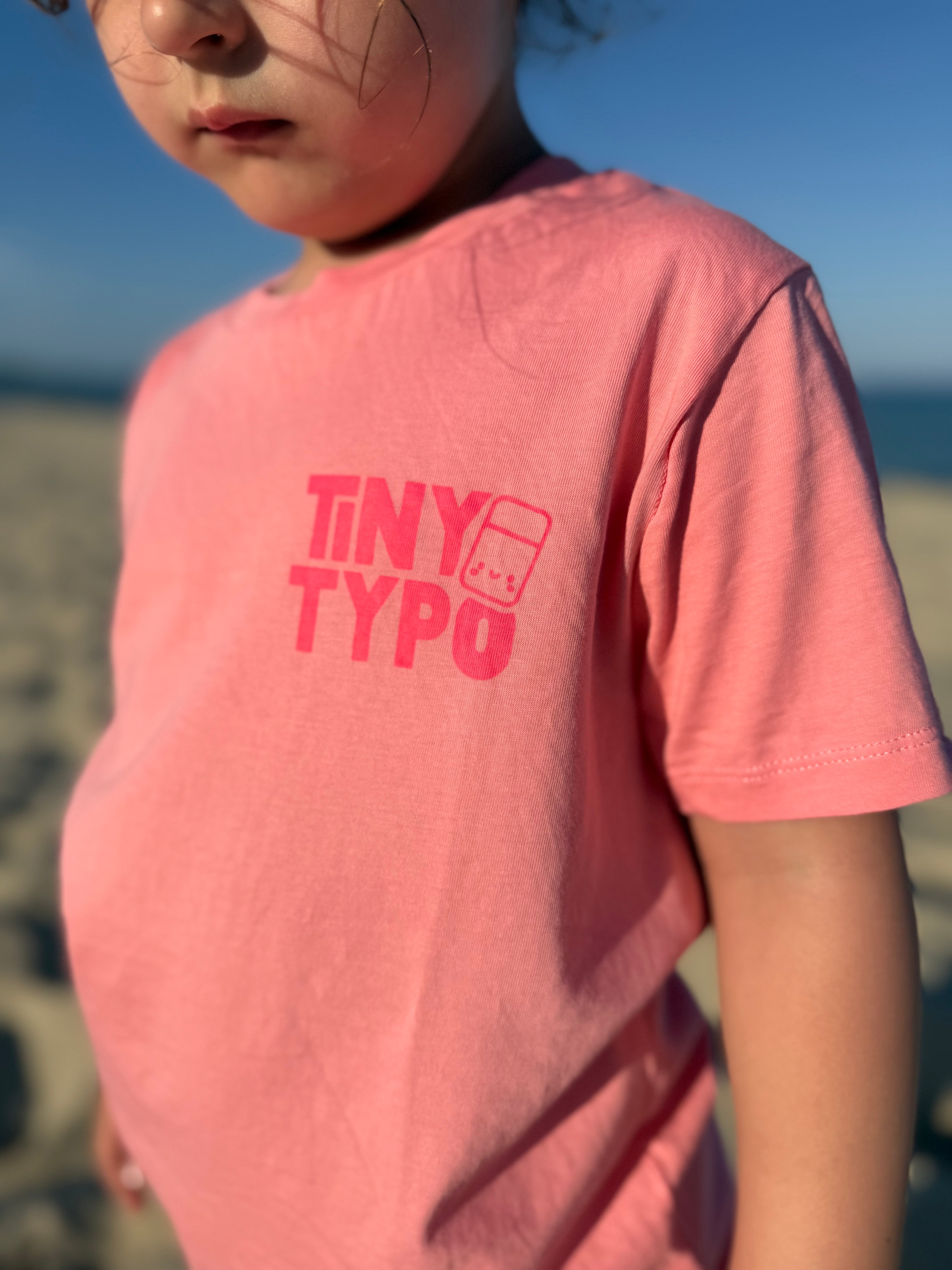 Kind trägt rosa Tiny-Typo T-Shirt mit Tiny Typo Logo am Strand – handbedruckt, nachhaltig