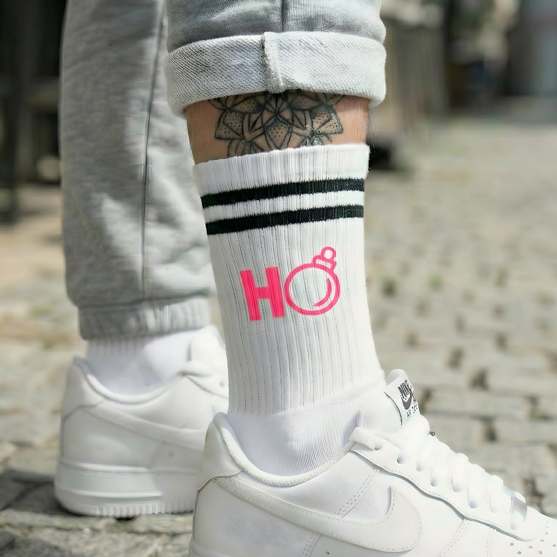 Weiße Socke mit pinkem „Ho“ Weihnachtsmotiv, getragen zu weißen Sneakern; urbaner Look auf Kopfsteinpflaster.