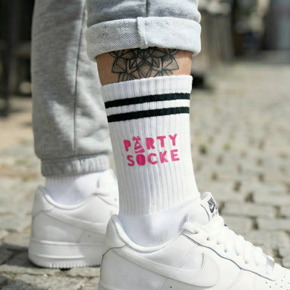 Weiße Socke mit pinkem „Party Socke“ Weihnachtsmotiv, getragen zu weißen Sneakern; urbaner Look auf Kopfsteinpflaster.