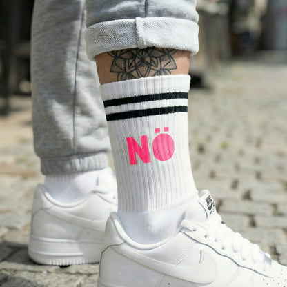 Weiße Socke mit pinkem „NÖ“ Statement-Print, getragen zu weißen Sneakern im Streetstyle auf Kopfsteinpflaster.