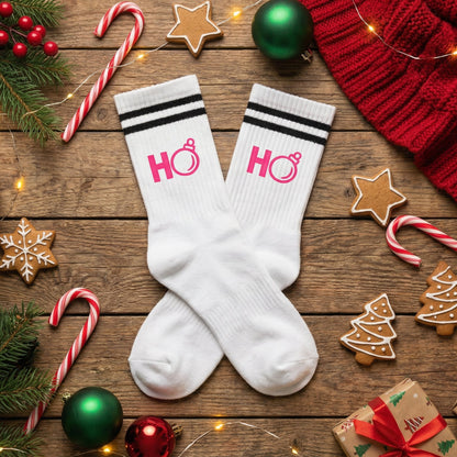 Weiße TinyTypo Socke mit pinkem „Ho Ho“ Print und Partyhut-Icon, dekoriert zwischen Weihnachtskugeln, Tannenzweigen, Geschenken und Kakao.
