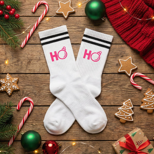 Weiße TinyTypo Socke mit pinkem „Ho Ho“ Print und Partyhut-Icon, dekoriert zwischen Weihnachtskugeln, Tannenzweigen, Geschenken und Kakao.