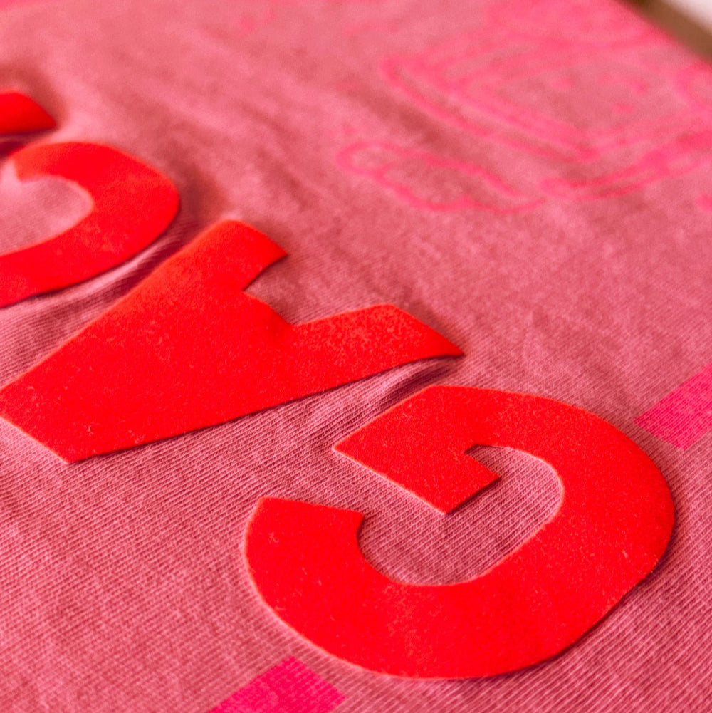 Detailaufnahme eines pinken T-Shirts mit neonorangenen, aus Velour geschnittenen Buchstaben, die auf den Stoff gepresst werden.