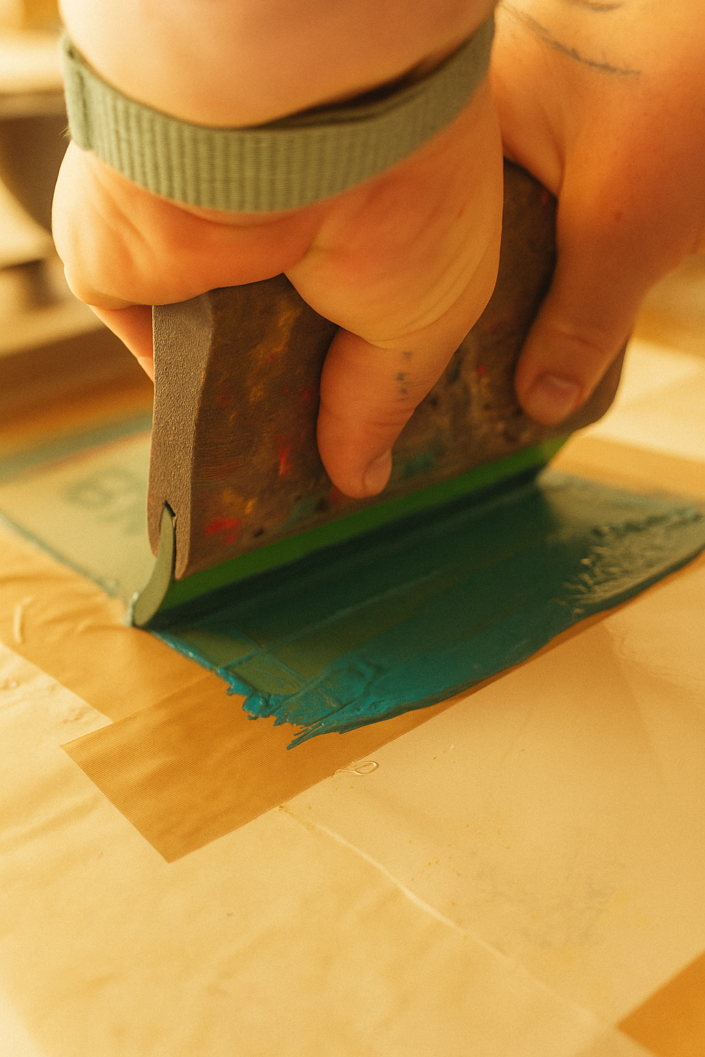 Nahaufnahme von Händen beim Siebdruck, wie ein Rakel türkisfarbene Farbe über ein mit Klebeband fixiertes Sieb zieht – in warmem Licht, das Handwerk und Detailreichtum betont.